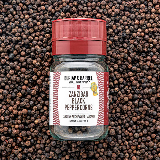 A bottle of Burlap & Barrel Zanzibar Black Peppercorns centered against a dense background of whole black peppercorns. The bottle has a red screw-top lid and a white label with red side accents.