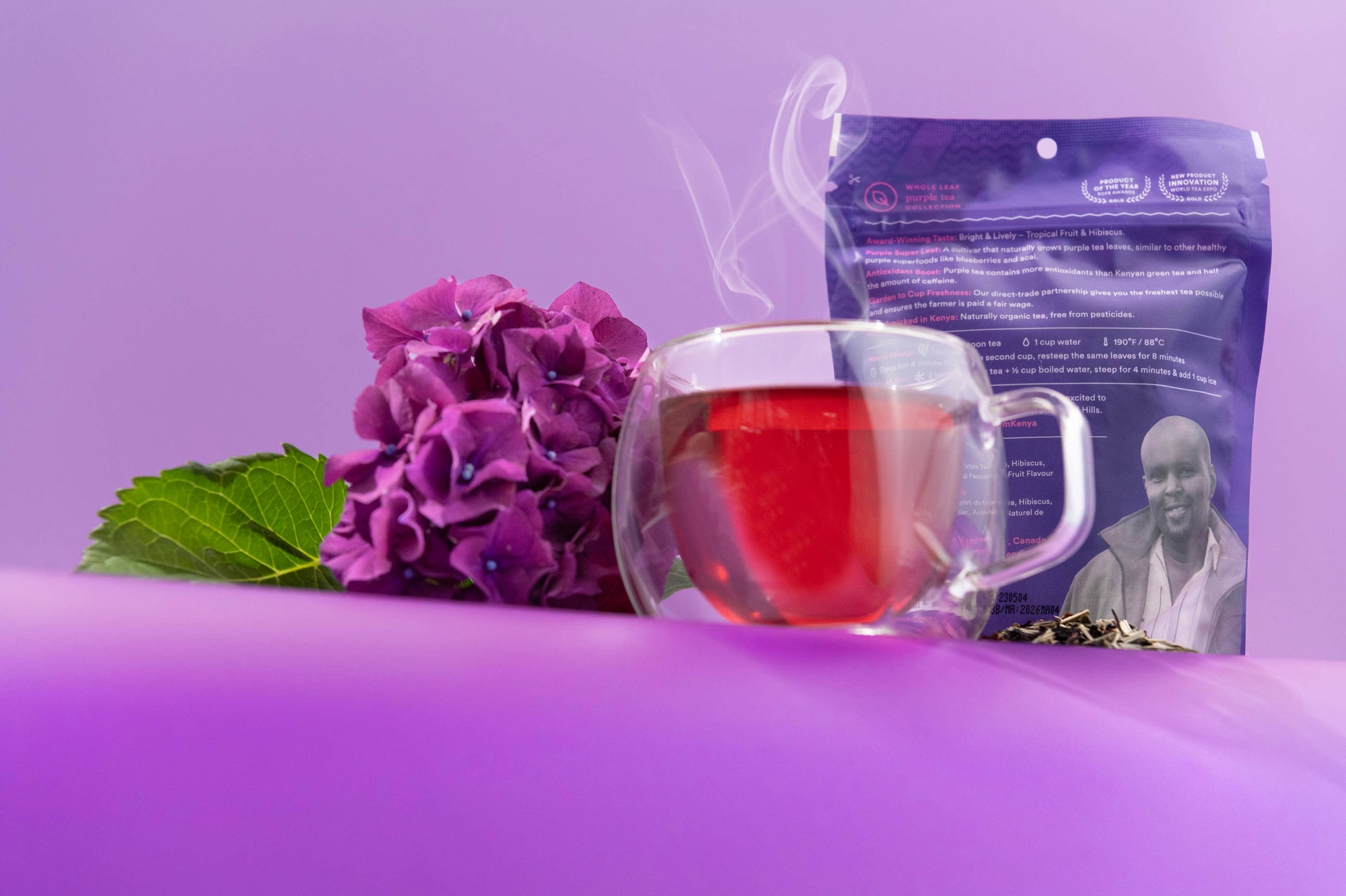 Tea bag with a purple package featuring a person, a glass of red tea, and purple flowers on a purple background.