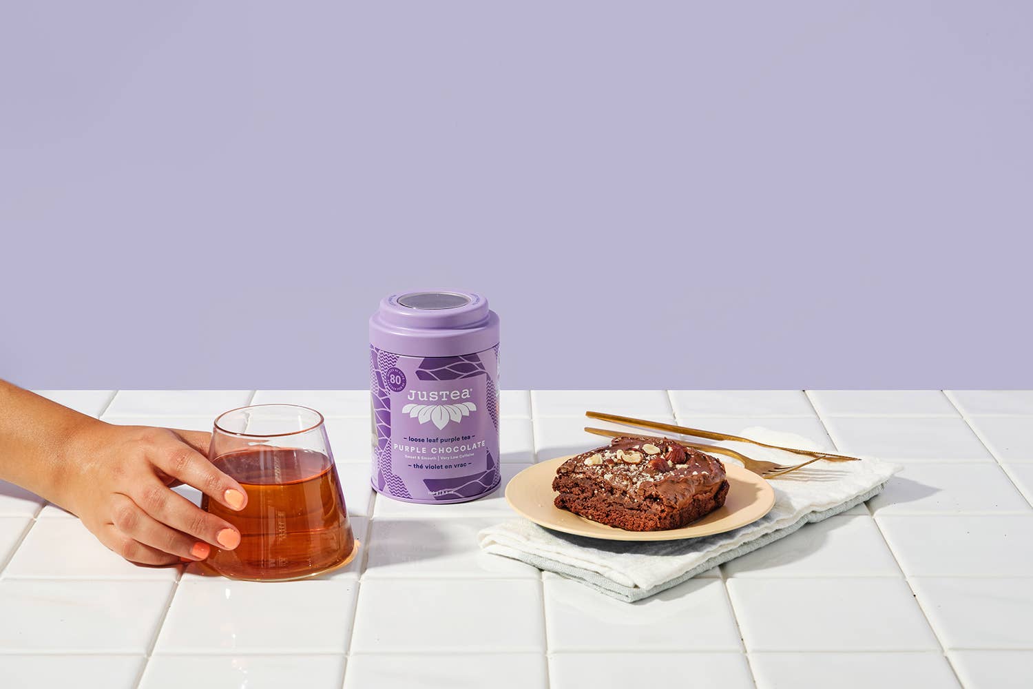 Person holding a glass of tea next to a container of JusTea purple chocolate tea and a brownie on a white surface with a lavender purple background.