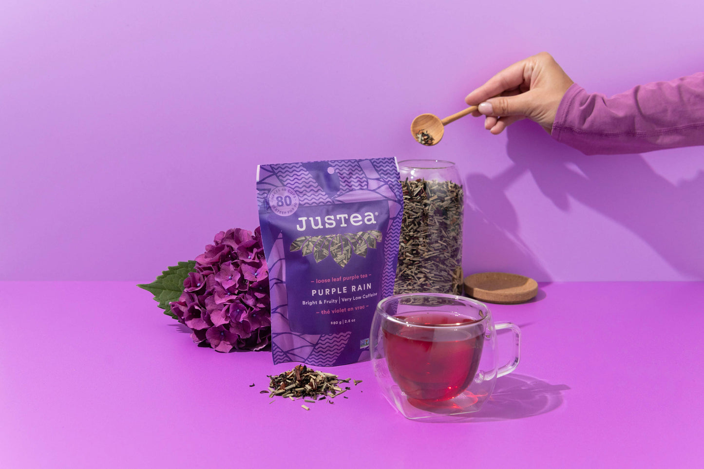 Person preparing a cup of Justea Purple Rain tea with a purple background