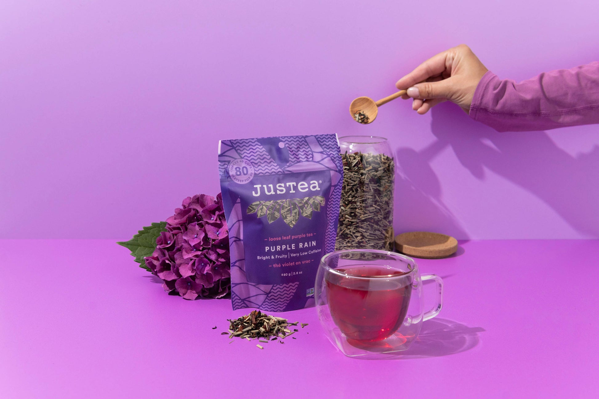 Person preparing a cup of Justea Purple Rain tea with a purple background