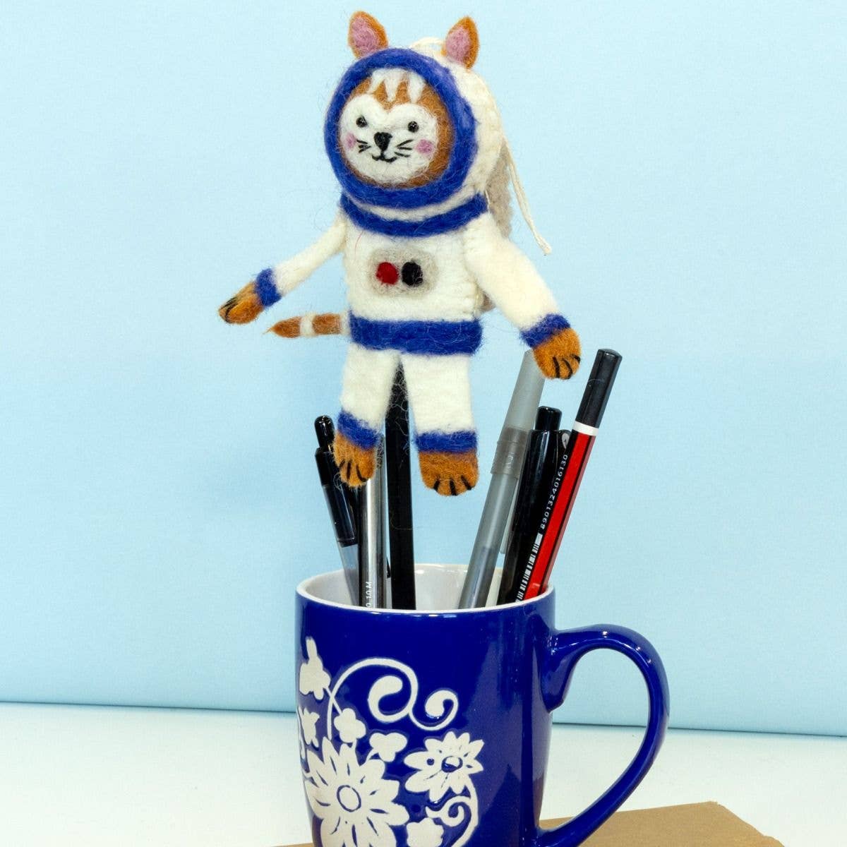 A felt cat astronaut toy standing upright in a blue ceramic mug filled with pens, against a light blue background.