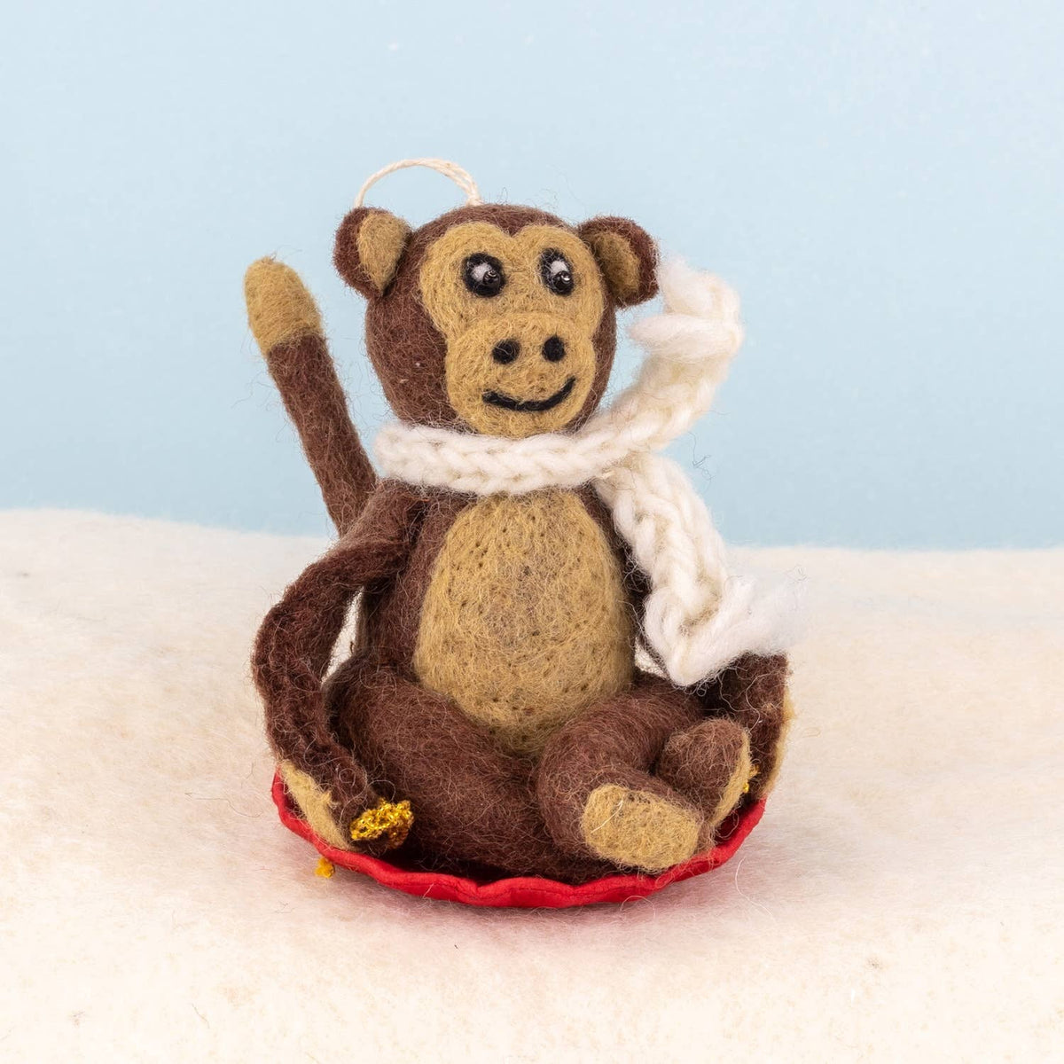 A felt monkey ornament wearing a white knit scarf and sitting on a red saucer, photographed against a soft light blue background with a white textured surface below.