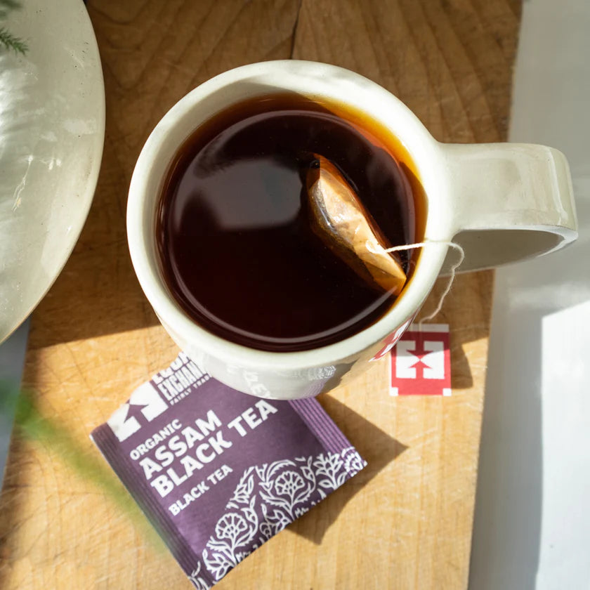 White mug with Assam black tea on a wooden surface, next to a tea bag and packaging.