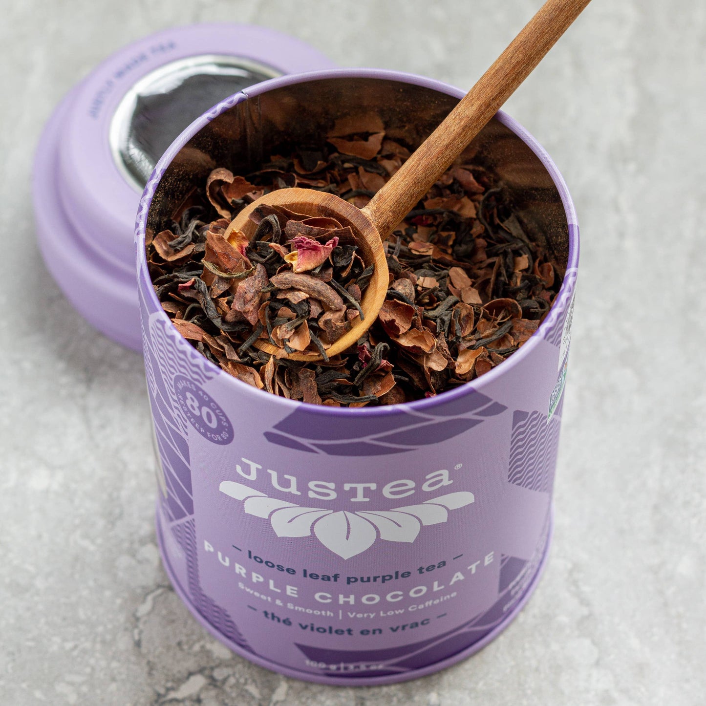 Purple container of Justea purple chocolate tea with a wooden spoon on a light gray background