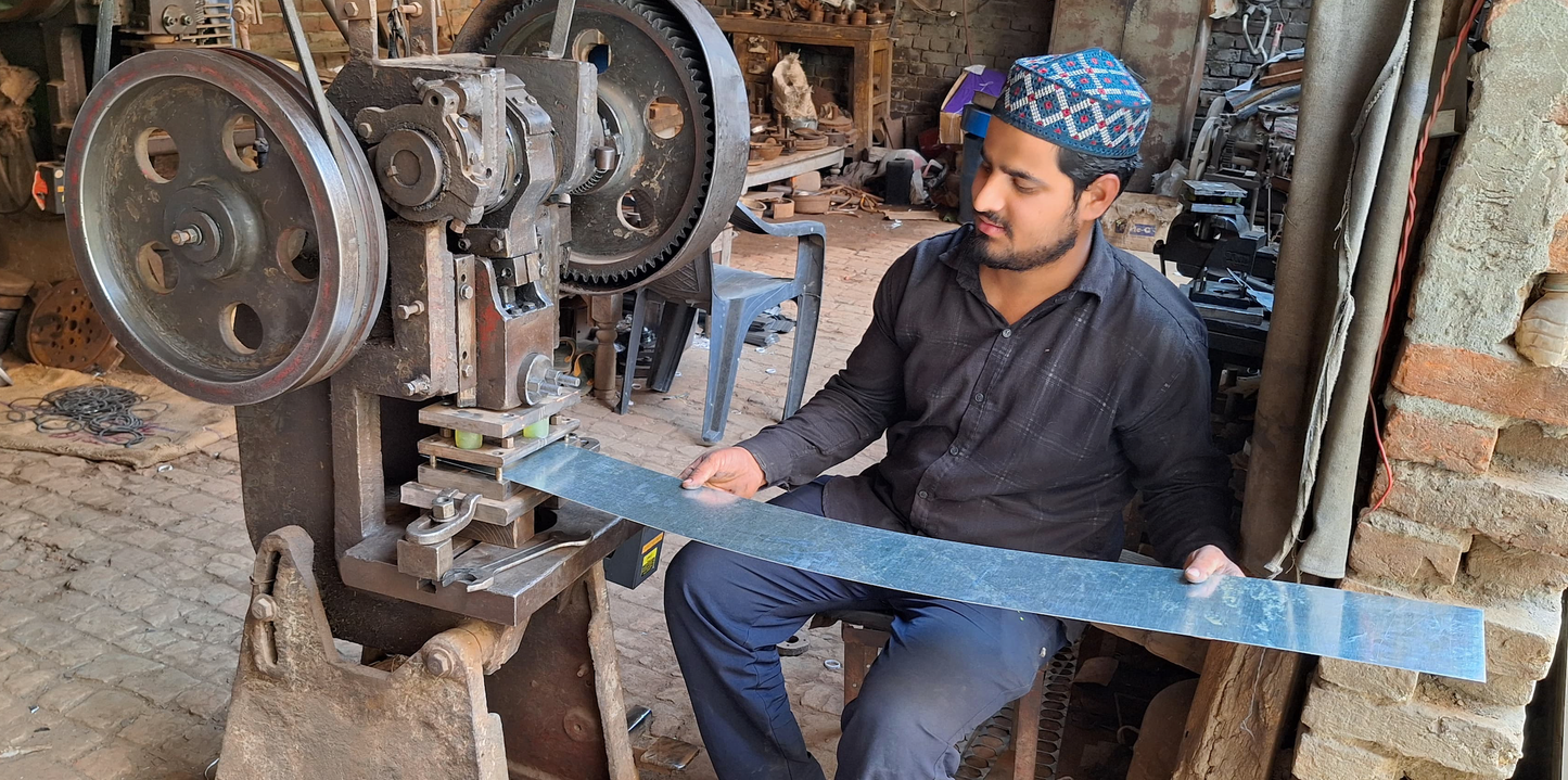 Man feeding a sheet of stainless steel through a metal stamping machine that will cut out the pieces of metal that will become the tea strainer. Photo is set in a workshop with machinery and tools around.