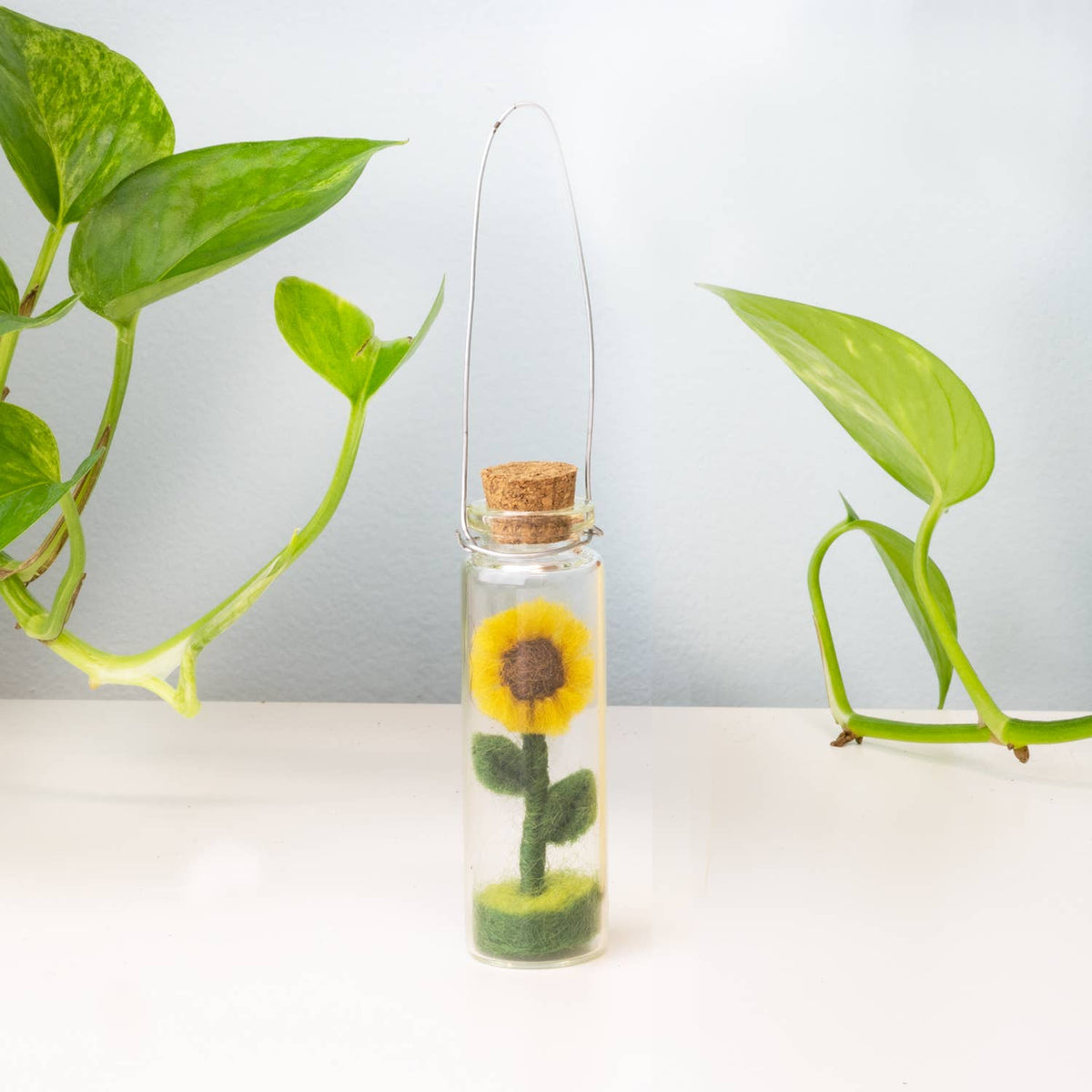 Small glass bottle with cork lid containing a felt sunflower against a light background with green leaves.