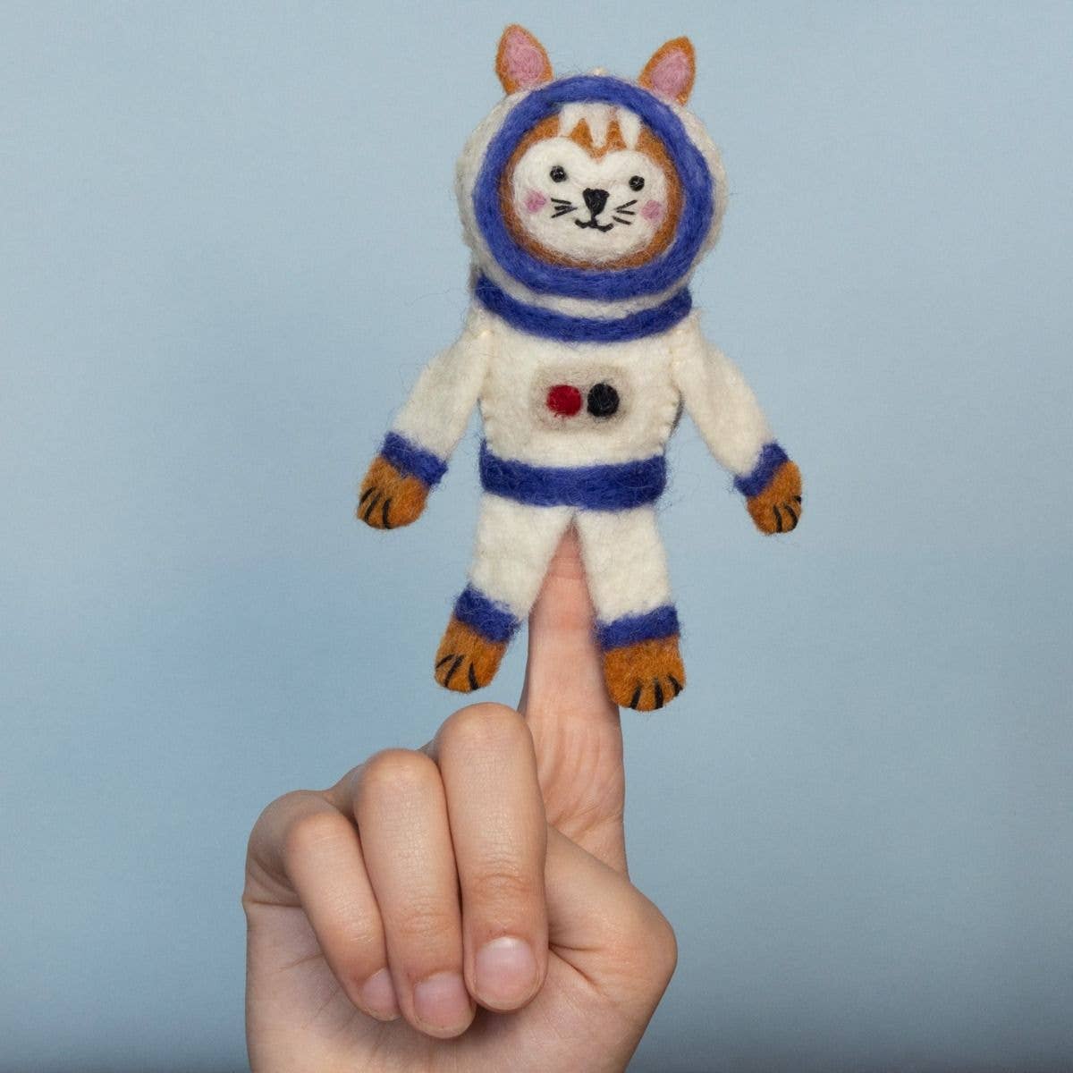 A small felt cat figurine dressed in a blue and white astronaut suit, held on a person's index finger against a soft light blue background.
