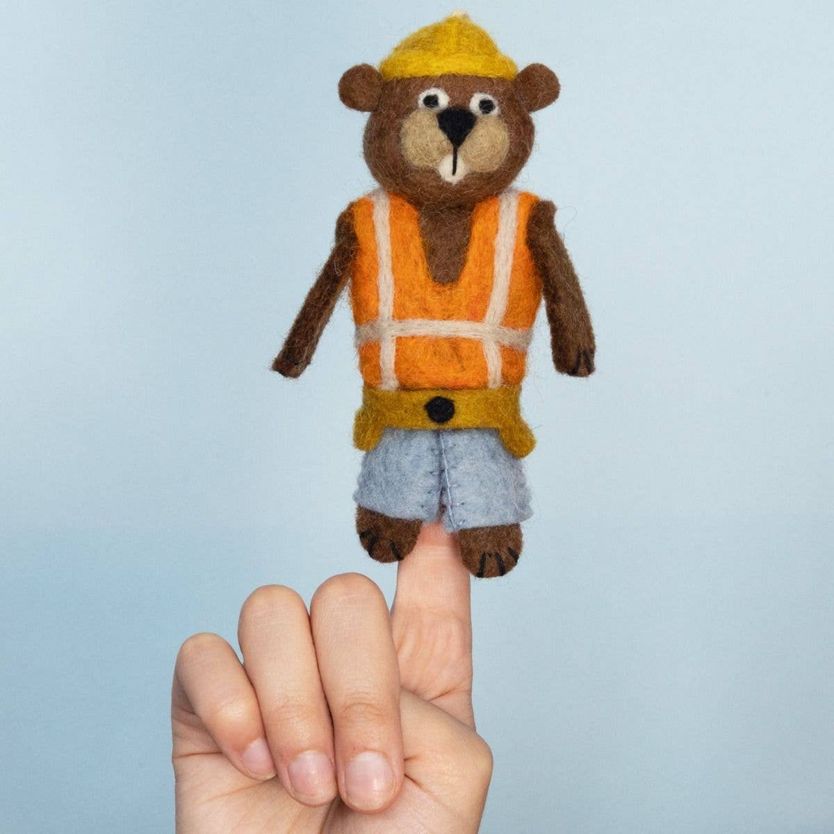 A small felted wool construction beaver figurine with a yellow hard hat, orange safety vest, and blue shorts, shown resting on a person's finger against a light blue background.