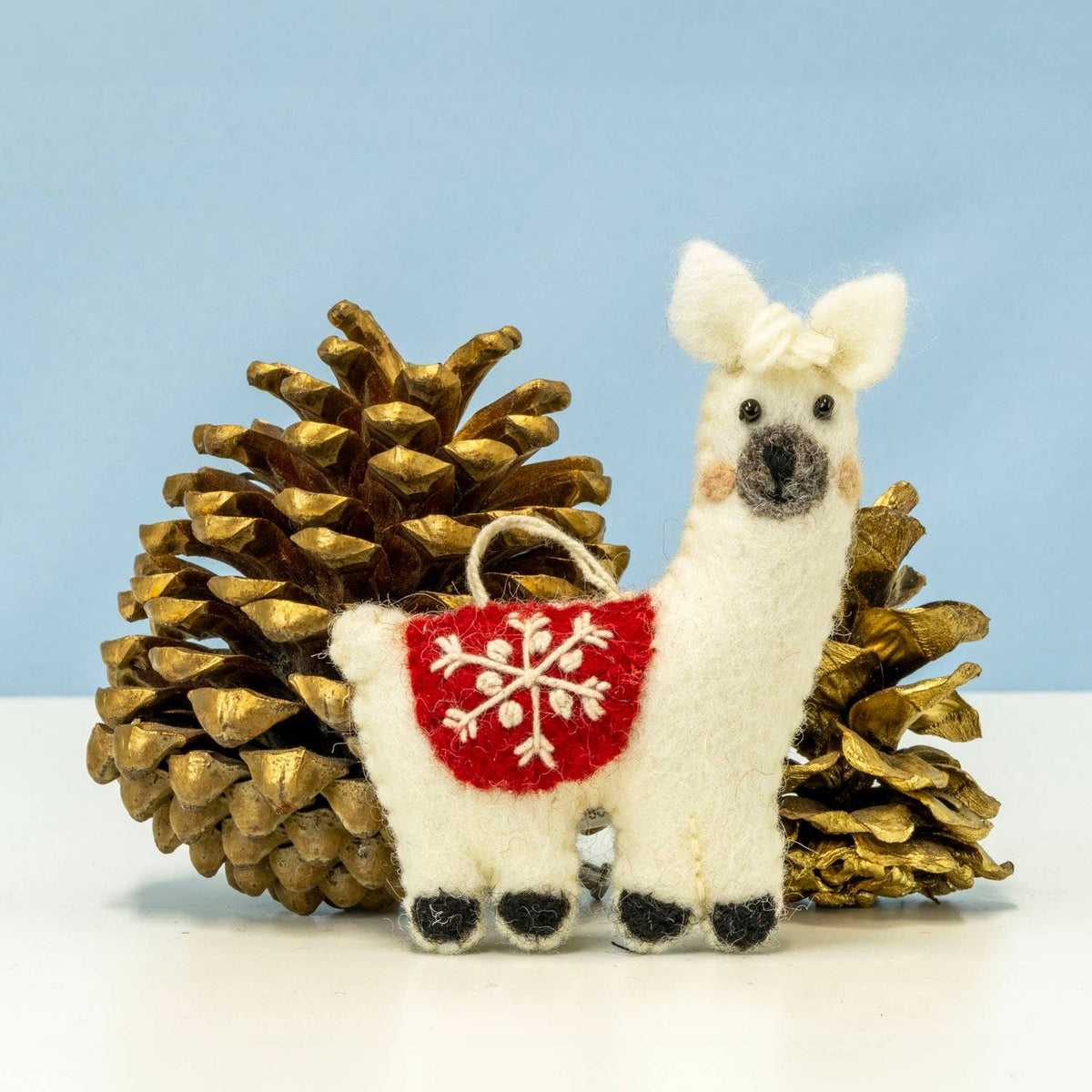 A white felt llama ornament with a red belly patch featuring a white snowflake design, posed between two gold pinecones against a light blue background.