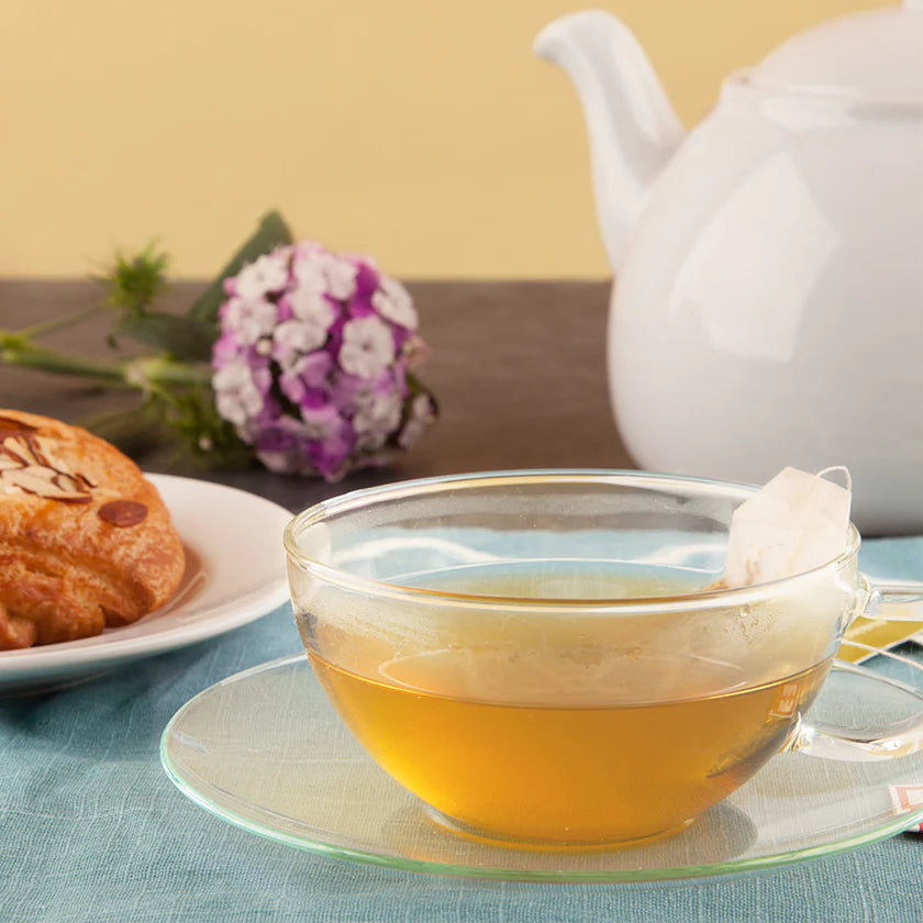 Clear glass mug with tea on a saucer, next to a white teapot and pastries on a table.