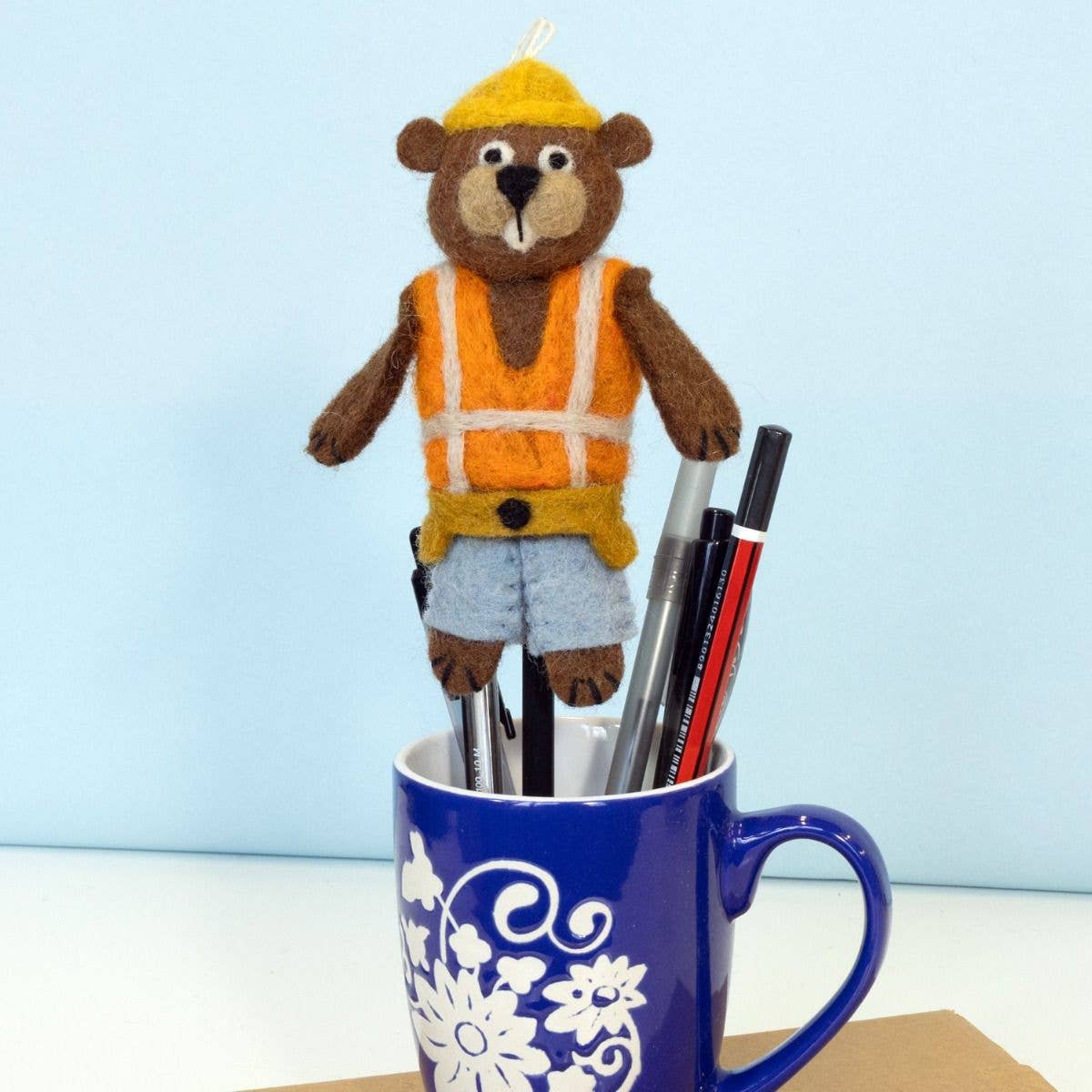 A felt beaver office accessory with a yellow hard hat, orange safety vest, and blue shorts, holding several pens in a blue mug with a white floral pattern against a light blue background.