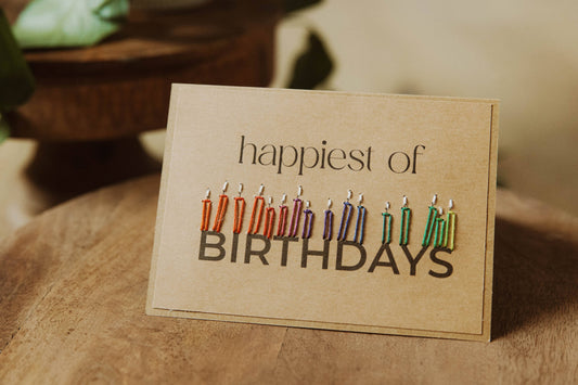 Greeting card with colorful candles and 'happiest of birthdays' text styled on a wooden surface.