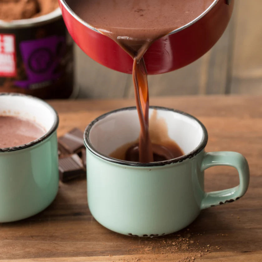 Hot chocolate being poured from a red pot into a green mug on a wooden surface.