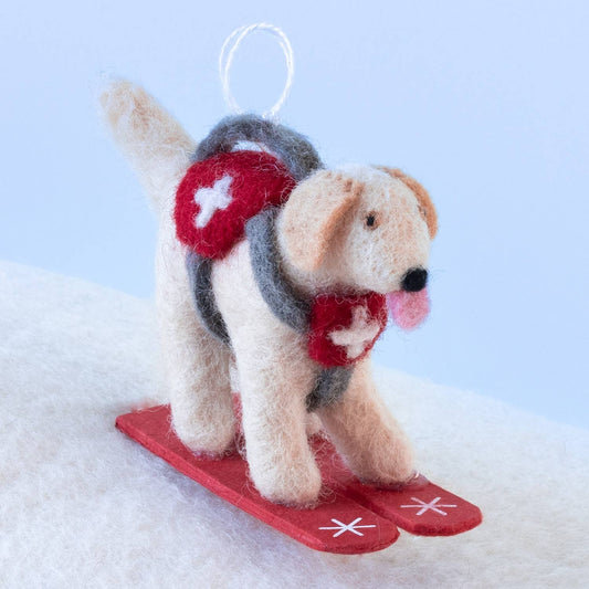 A felted wool dog ornament wearing a red and gray harness with white crosses, standing on red skis against a light blue and white background.