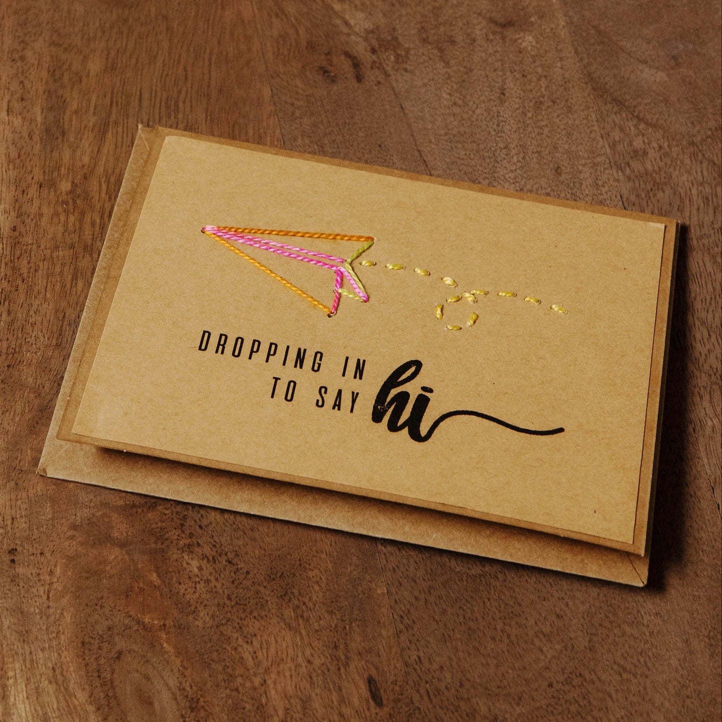 Brown greeting card with 'dropping in to say hi' text on a brown recycled paper card with a stitched paper airplane design.
