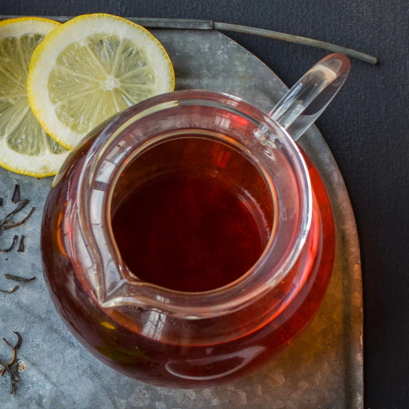 Glass teapot filled with red tea on a metal tray with lemon slices and tea leaves.