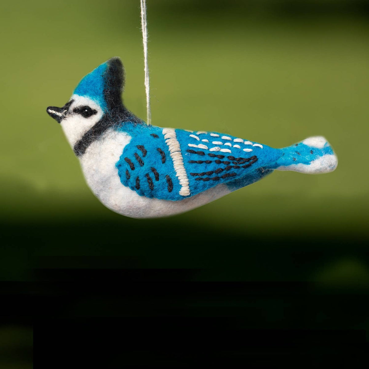 A felt bird ornament, likely a jay or bluebird, hanging by a thin string. The bird features bright blue, white, and black felted wool with embroidered details on its wings and back, set against a blurred green background.