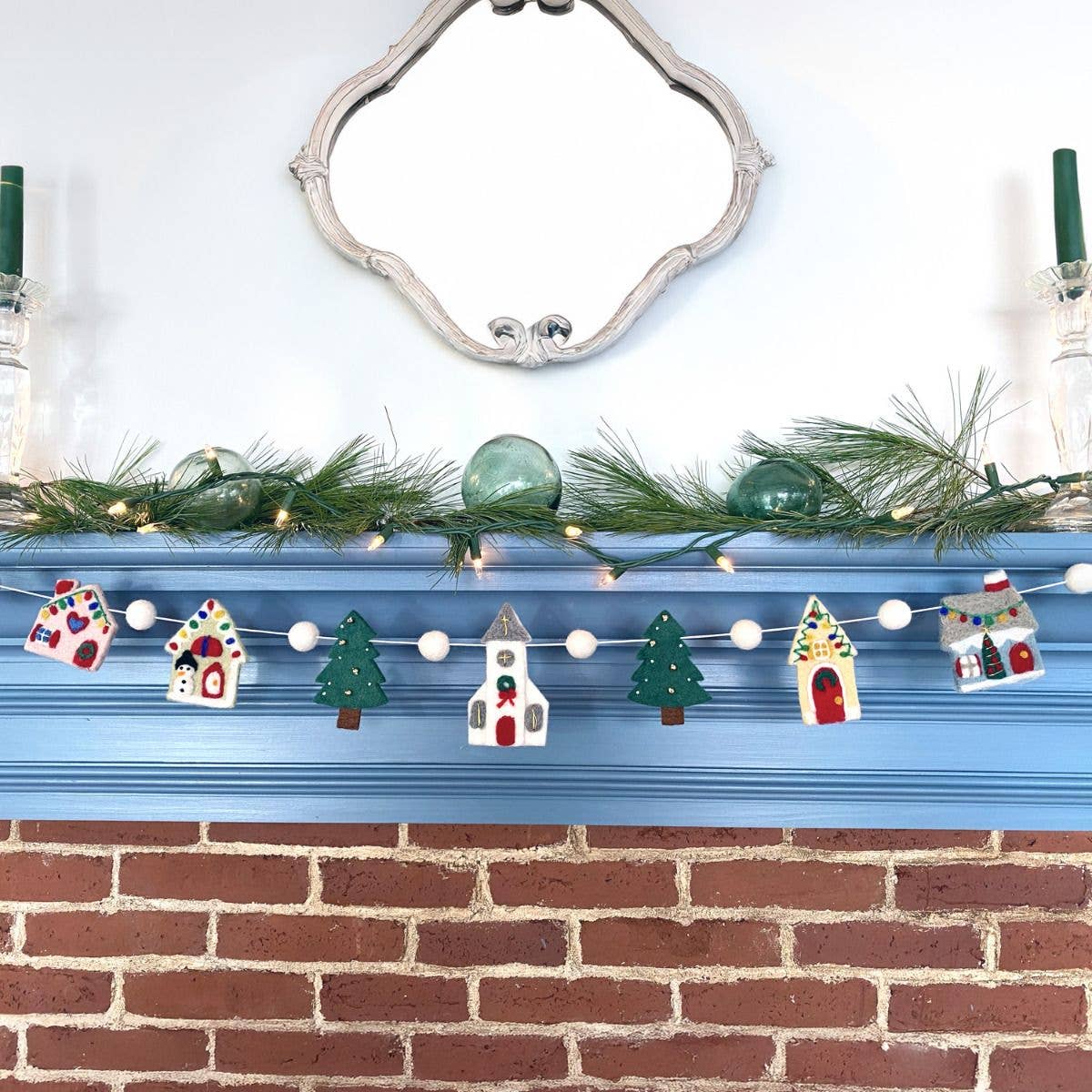 A festive Christmas garland featuring miniature white houses, green Christmas trees, and white beads draped over a blue fireplace mantel, with a brick fireplace below and a mirror above.