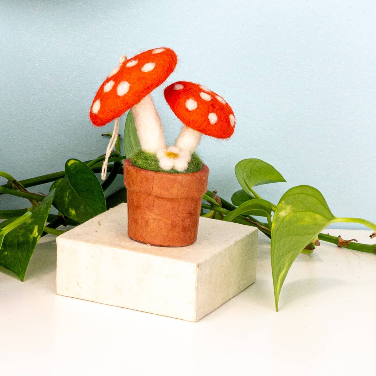 A small felted mushroom decoration in a terracotta pot with green grass and a white flower, displayed on a white block with green leaves against a light blue background.
