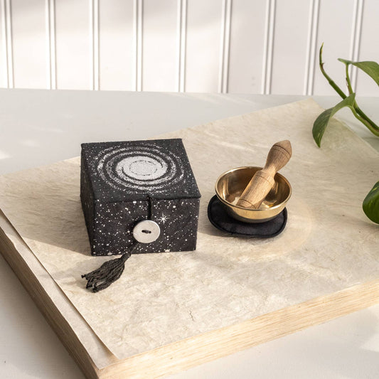 Black decorative box with white patterns and a brass singing bowl with a wooden striker on a stack of papers on a wooden  surface