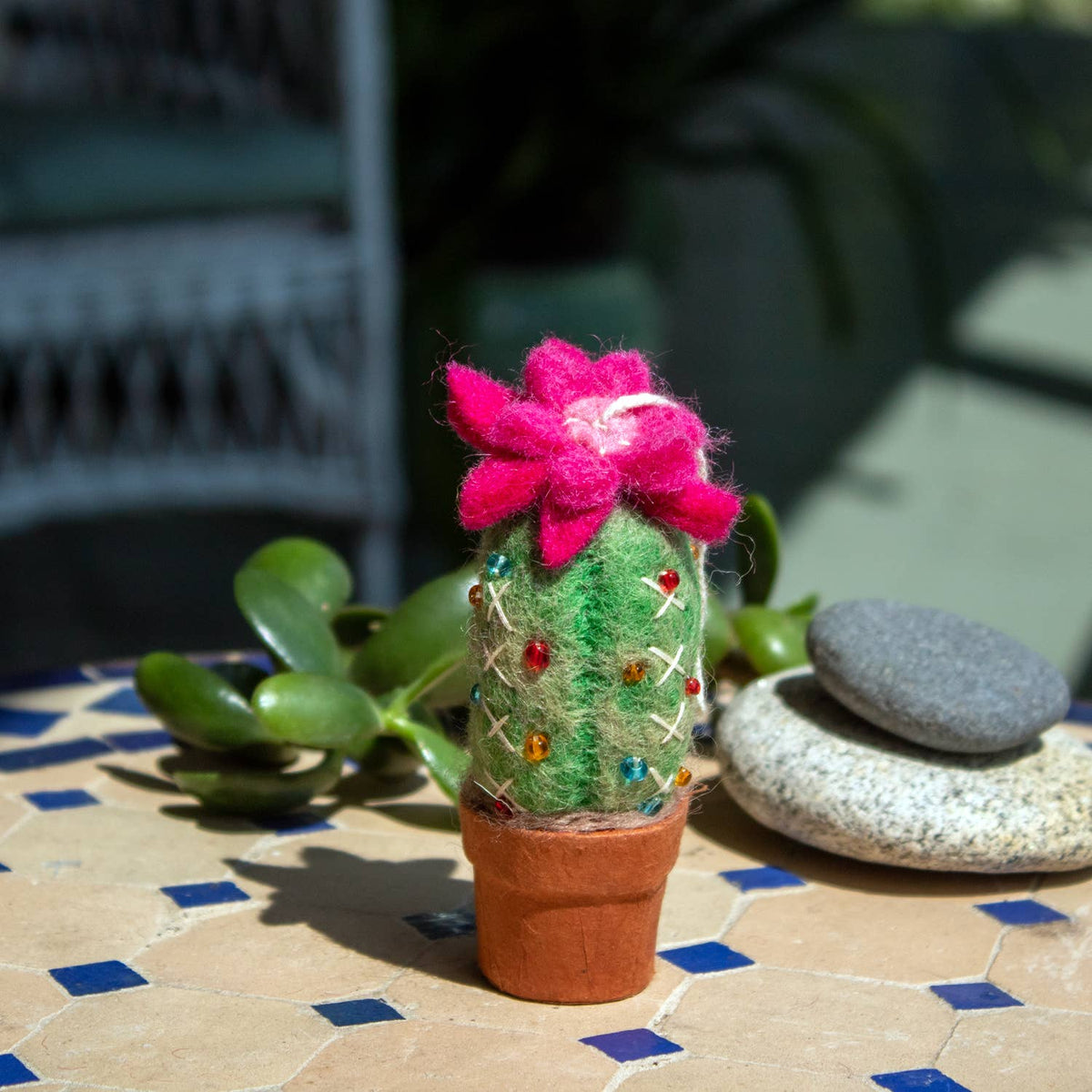 A felted wool cactus decoration in a terracotta pot, decorated with a bright pink felt flower and colorful rhinestones, set on a tiled patio next to a succulent.