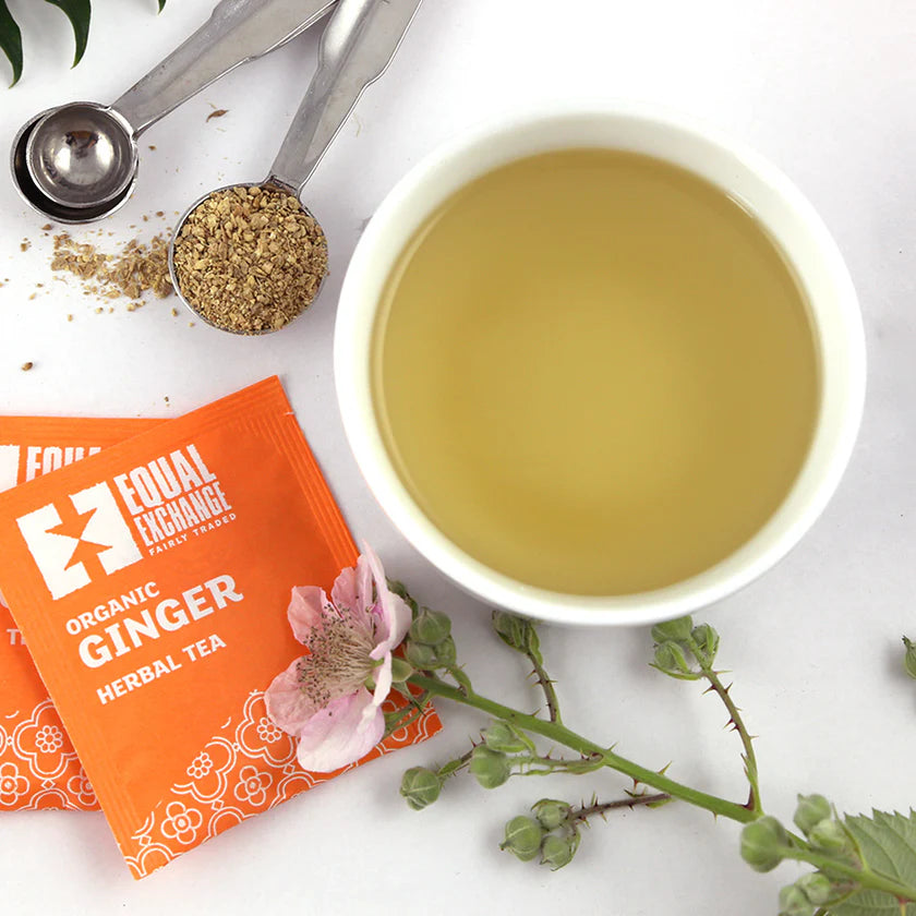White cup of tea with Equal Exchange organic ginger herbal tea packets, dried ginger, and flowers on a white background.