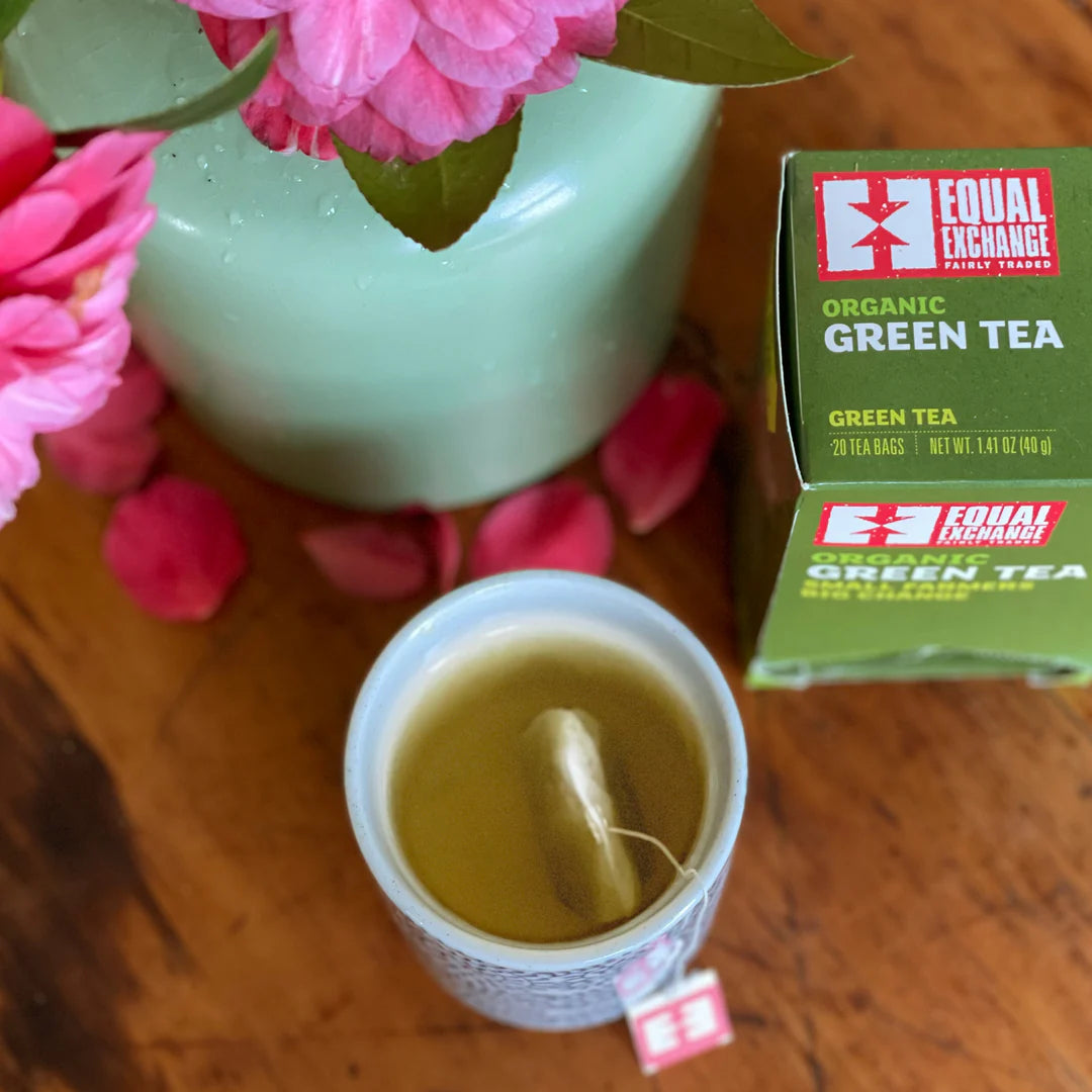 Green tea cup with a tea bag, pink flowers, and Equal Exchange green tea box on a wooden surface.