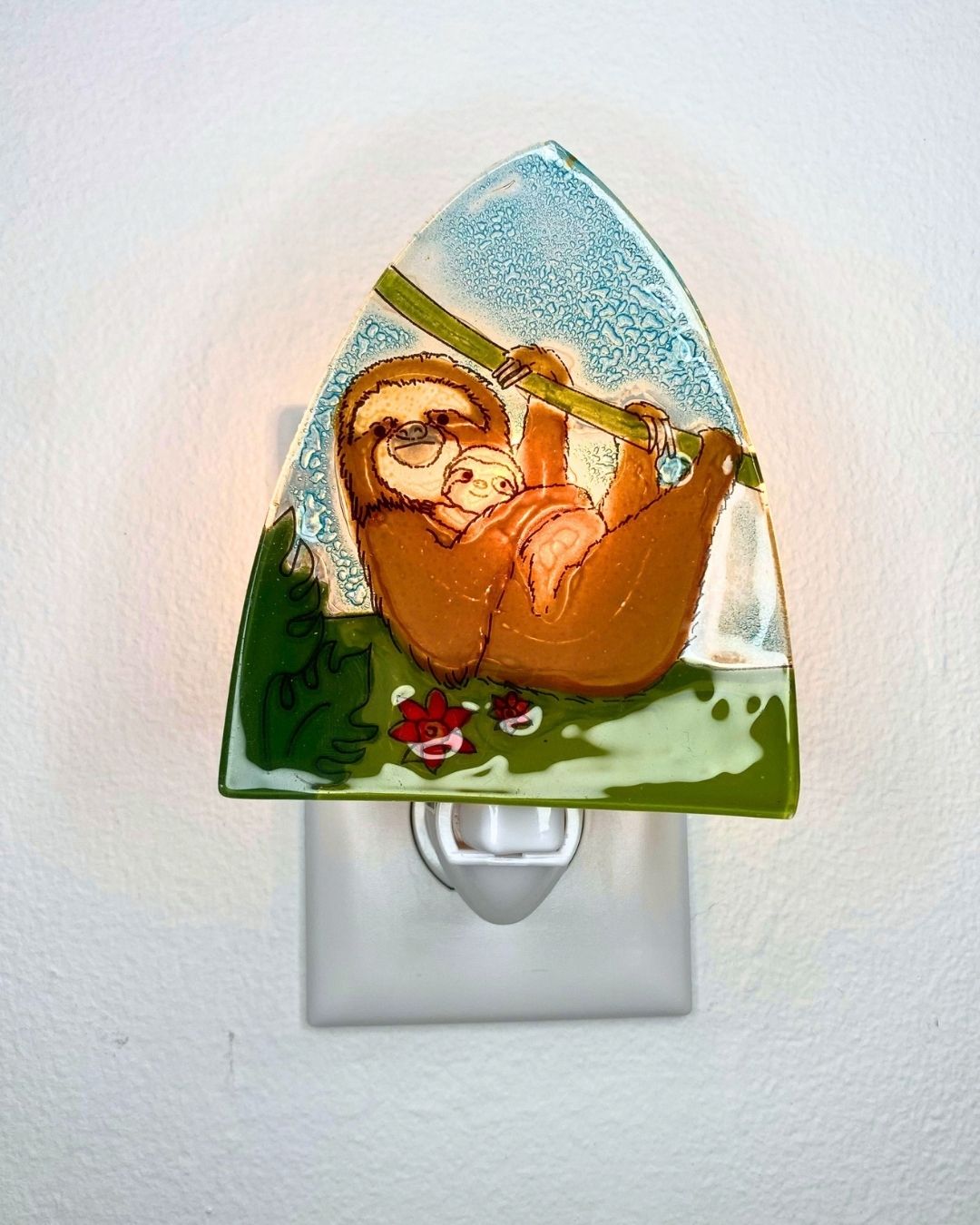 A decorative triangular-shaped night light featuring a hand-painted illustration of a mother sloth holding a small baby sloth mounted on a white outlet against a white wall. 
