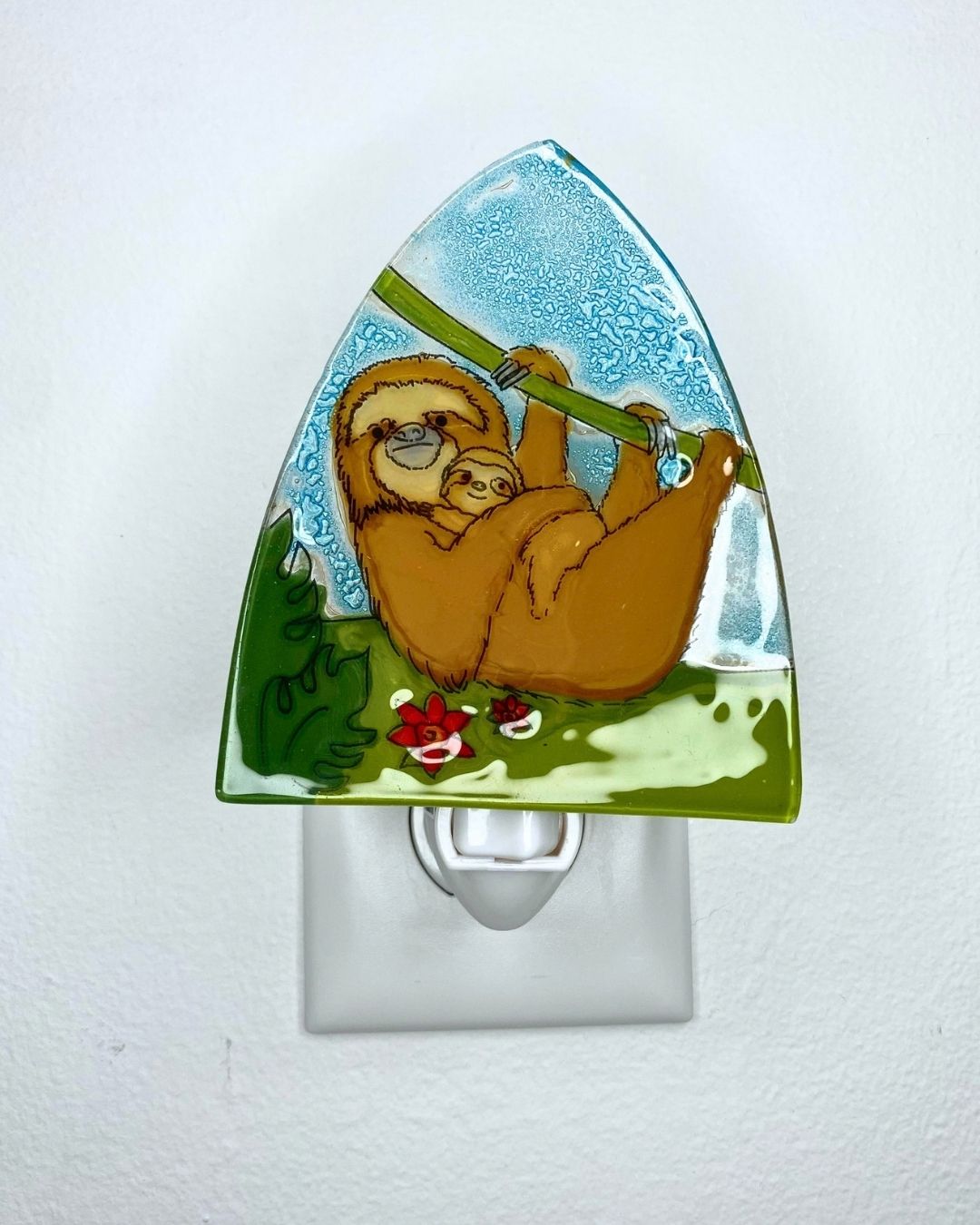 A decorative triangular-shaped night light featuring a hand-painted illustration of a mother sloth holding a small baby sloth mounted on a white outlet against a white wall.