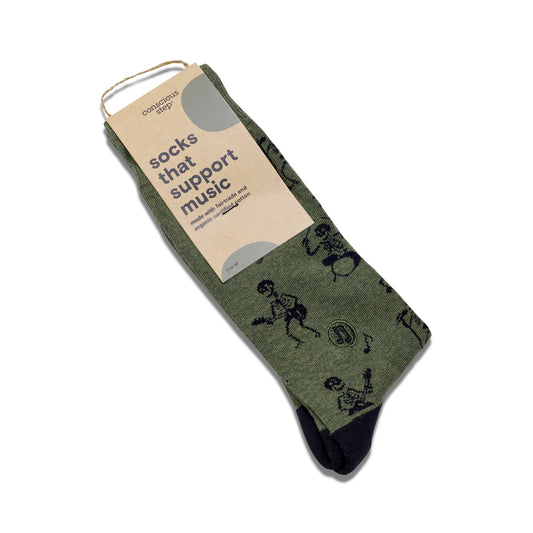 A pair of green crew socks with a black skeleton pattern, packaged with a cardboard tag that reads 'socks that support music' on a white background.