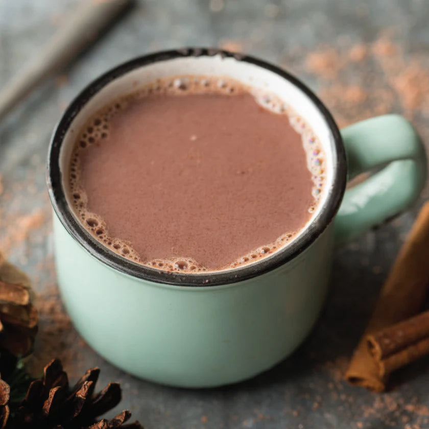 Teal mug filled with hot chocolate on a rustic surface with cinnamon sticks and pine cones.