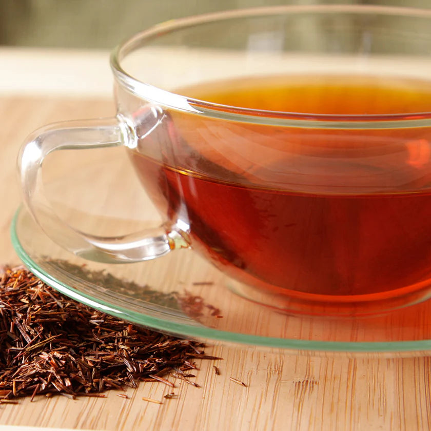 Clear glass cup filled with red vanilla rooibos tea on a wooden surface with loose tea leaves.